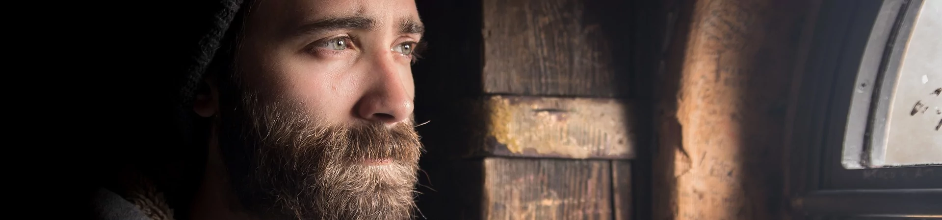 A bearded man in a hoodie gazes thoughtfully out of a window, lost in contemplation.