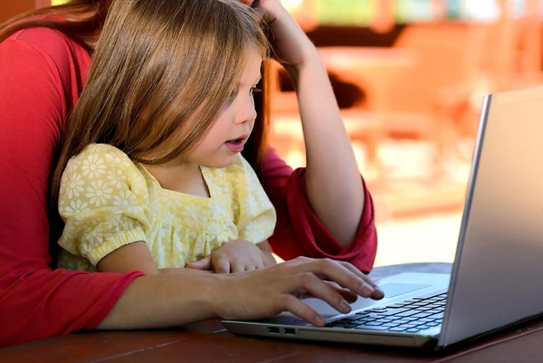 A woman and a child sit together at a table, engaged with a laptop between them.
